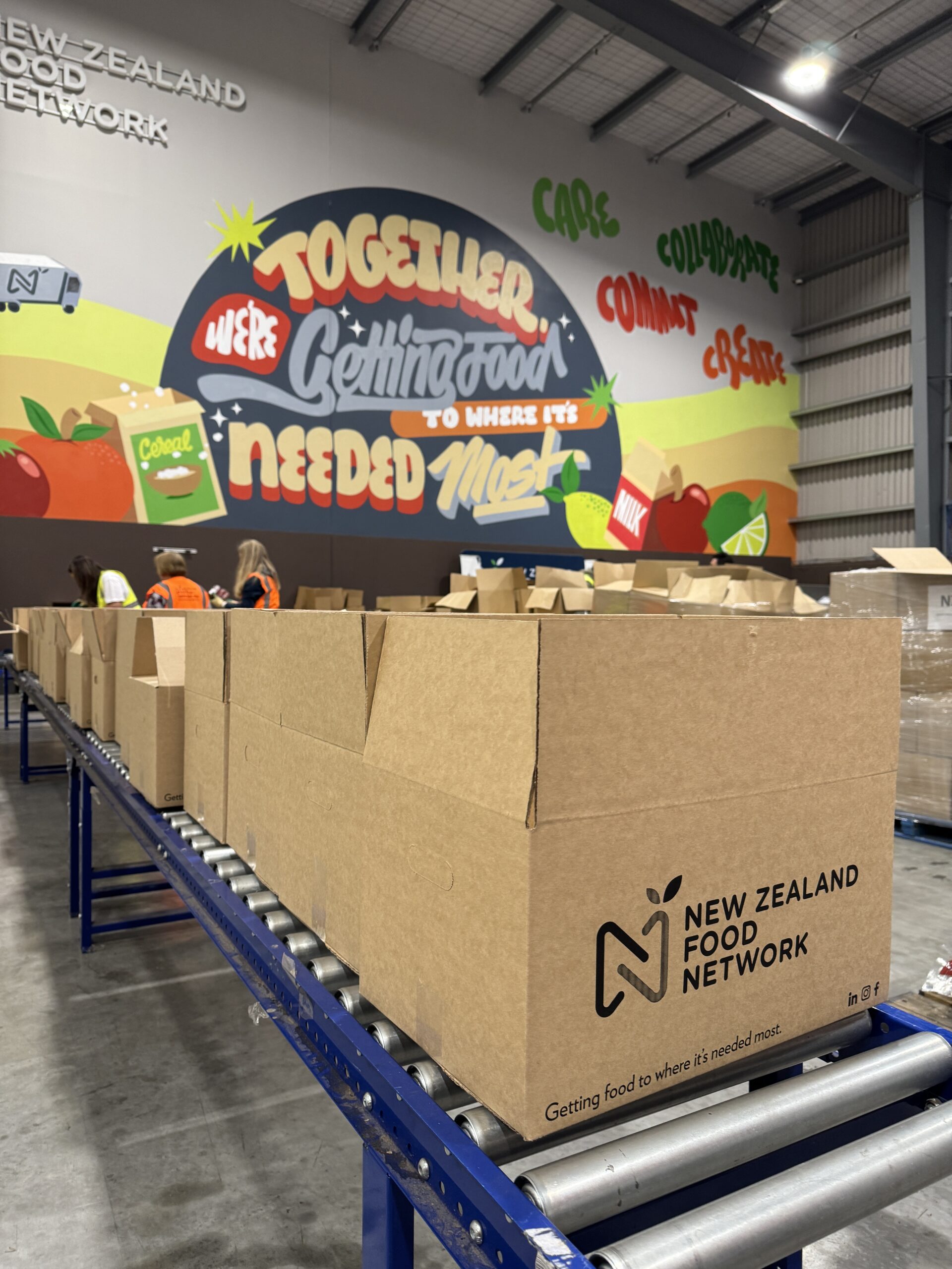 Food boxes on a conveyor in a New Zealand Food Network warehouse beneath a mural about getting food to where it’s needed most.