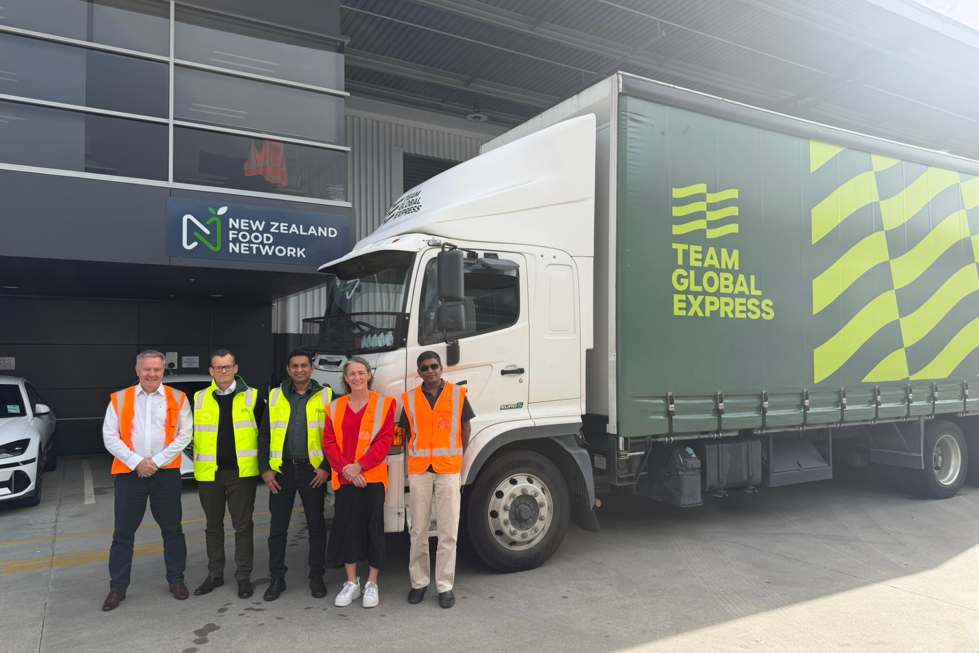 Food distribution in NZFN logistics partnership with Team Global Express. A group standing beside a Team Global Express delivery truck outside a New Zealand Food Network building.
