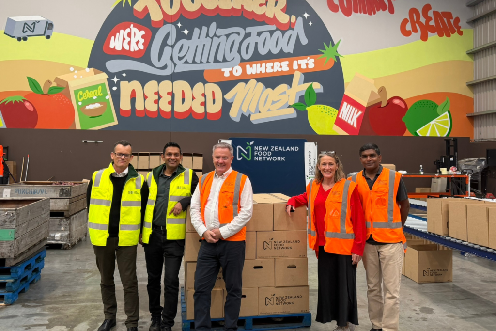 A group standing in a warehouse beside stacked NZ Food Network boxes, with a large mural reading “Getting food to where it’s needed most” in the background.