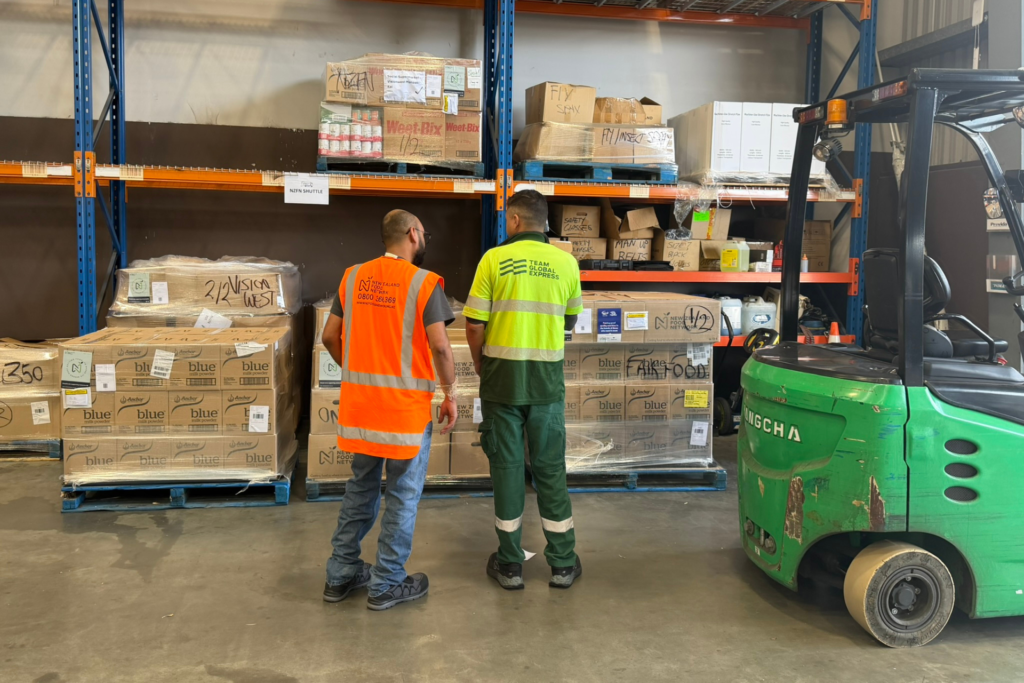 Two workers in high-visibility vests standing in a warehouse beside stacked pallets of boxed food, with shelving units and a forklift nearby.