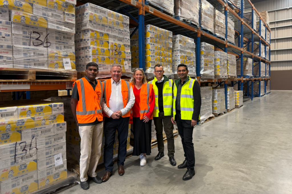 A group standing in a warehouse beside tall shelving filled with large pallets of boxed food supplies wrapped in plastic.